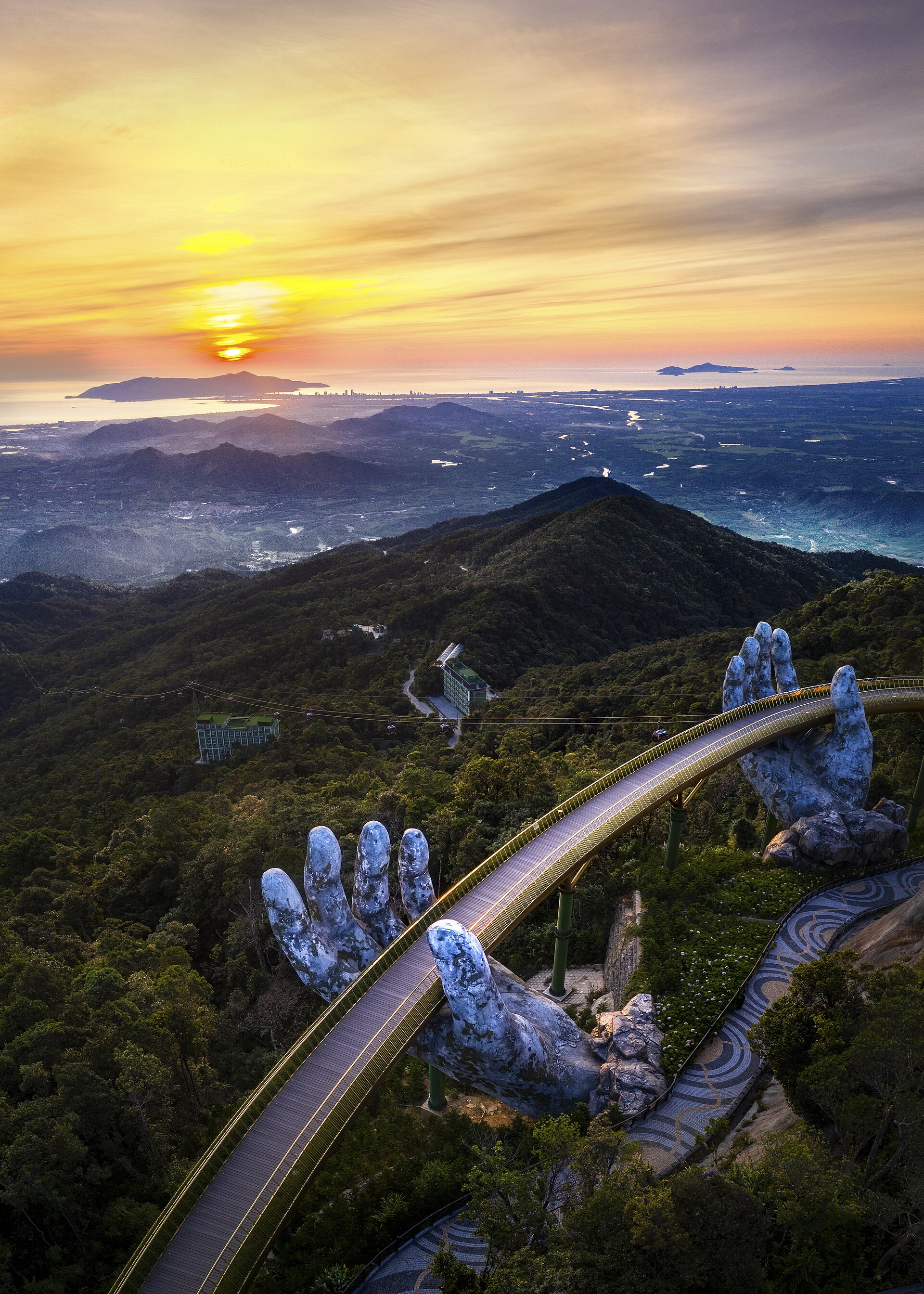 Aerial view of the Golden Bridge curving between two giant stone hands above forested hills at sunset